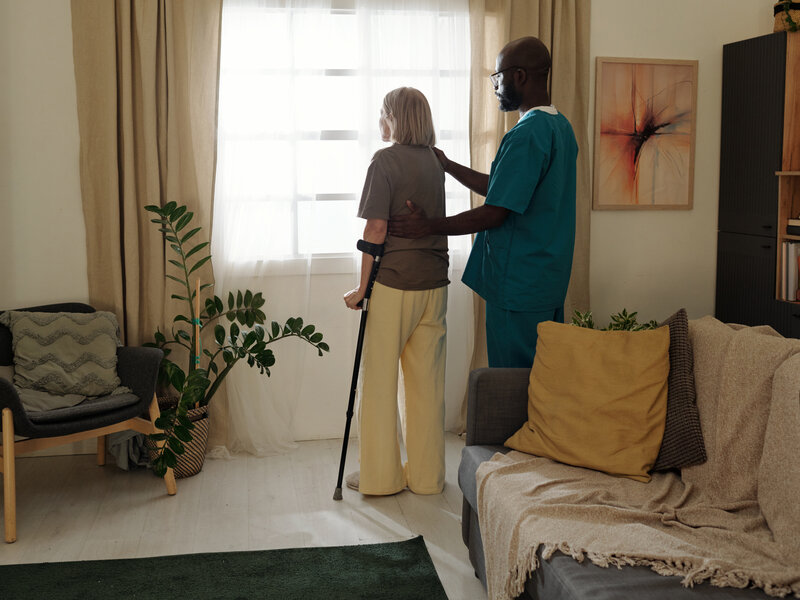Professional carer assisting elderly client with mobility support and walking aid in bright, comfortable living room