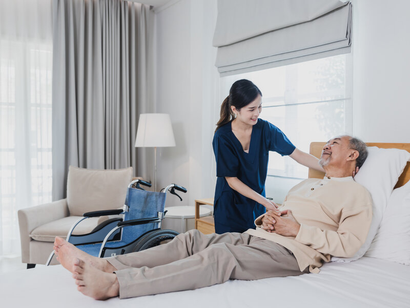 Professional female caregiver providing compassionate bedside care to senior Asian gentleman in comfortable modern healthcare environment