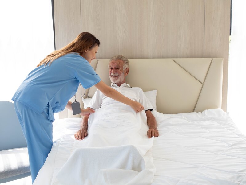 Professional female caregiver providing compassionate bedside care and comfort to elderly gentleman in modern, comfortable bedroom setting