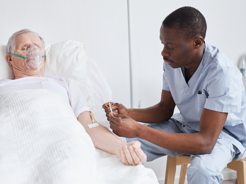 Professional male nurse administering IV therapy to elderly patient with oxygen support, demonstrating advanced clinical expertise and specialized medical care